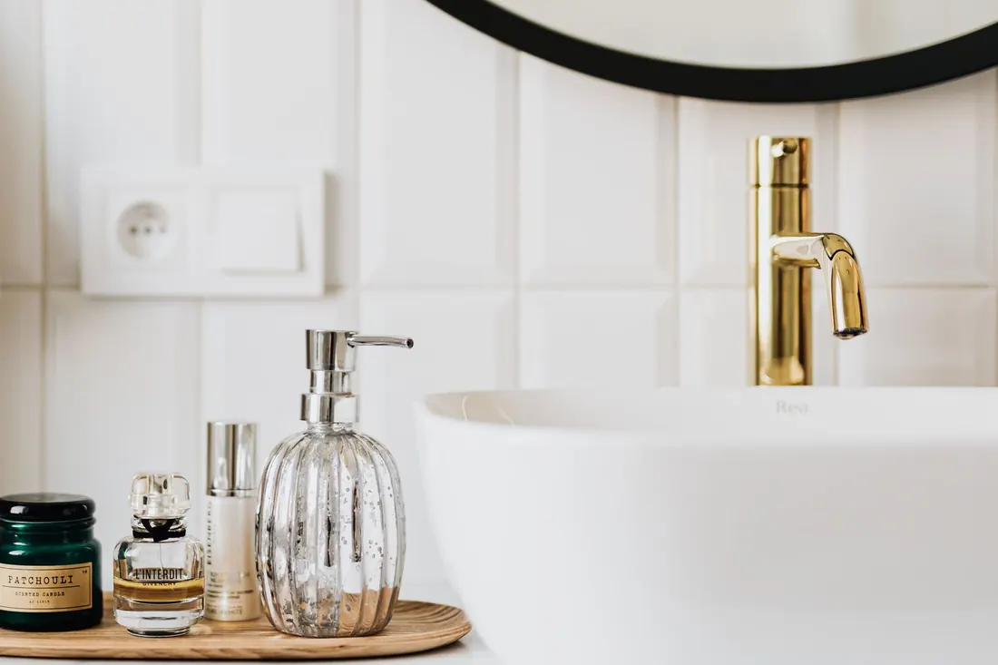 Bathroom vanity left spotless: a glass soap dispenser on a wooden tray beside a white basin and a polished brass tap